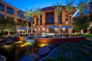 Modern courtyard with illuminated fountain, palm trees, patio seating, surrounded by office buildings at dusk.
