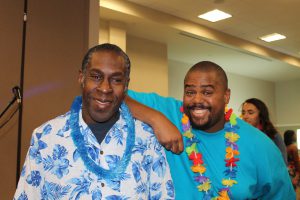 Two smiling men wearing colorful Hawaiian shirts and leis in a brightly lit room.