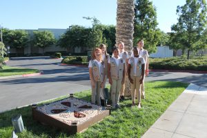 Group of six Girl Scouts in uniform standing near palm tree and circular garden.