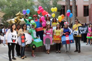 Group of people in colorful Mario Kart costumes holding balloons and signs, smiling outdoors.