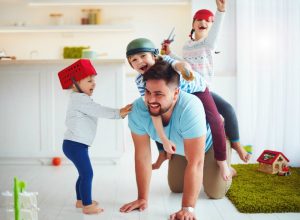 Father playing with three children wearing makeshift helmets, smiling and laughing in bright kitchen.