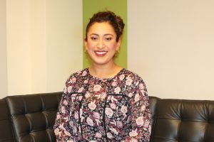 Smiling woman with floral dress sitting on black leather couch against green and beige wall.