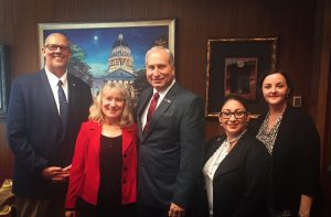 Five people standing together in office, framed painting of capitol building behind them.