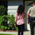 Man in uniform walking alongside woman towards white garage door of suburban home.