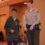 Boy wearing sheriff badge standing beside female officer and bloodhound in a building.