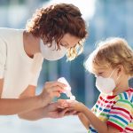 Woman and child in masks applying hand sanitizer indoors with bright natural light.