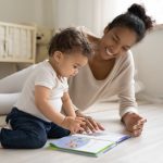 Mother and toddler reading colorful book on floor in bright room next to crib.