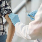Healthcare professional in blue gloves administering vaccine to person wearing plaid shirt.