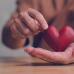 Hands holding and gently touching small red wooden heart on wooden table.