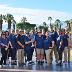 Group of people in blue shirts standing outdoors with palm trees and mountains in background.
