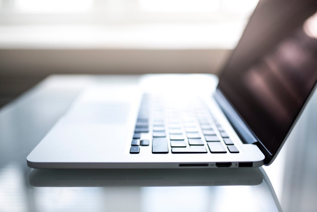 Open silver laptop on glass table, sunlight illuminating keyboard and touchpad, screen partially visible.
