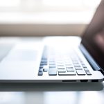 Open silver laptop on glass table, sunlight illuminating keyboard and touchpad, screen partially visible.