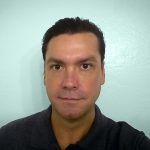 Man with dark hair and brown eyes against plain light blue background, wearing black shirt.