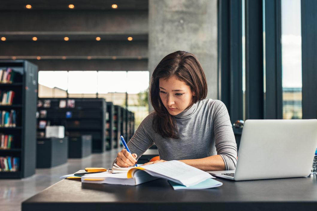 Woman in library studying with open book, taking notes, laptop nearby on table.