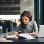 Woman in library studying with open book, taking notes, laptop nearby on table.