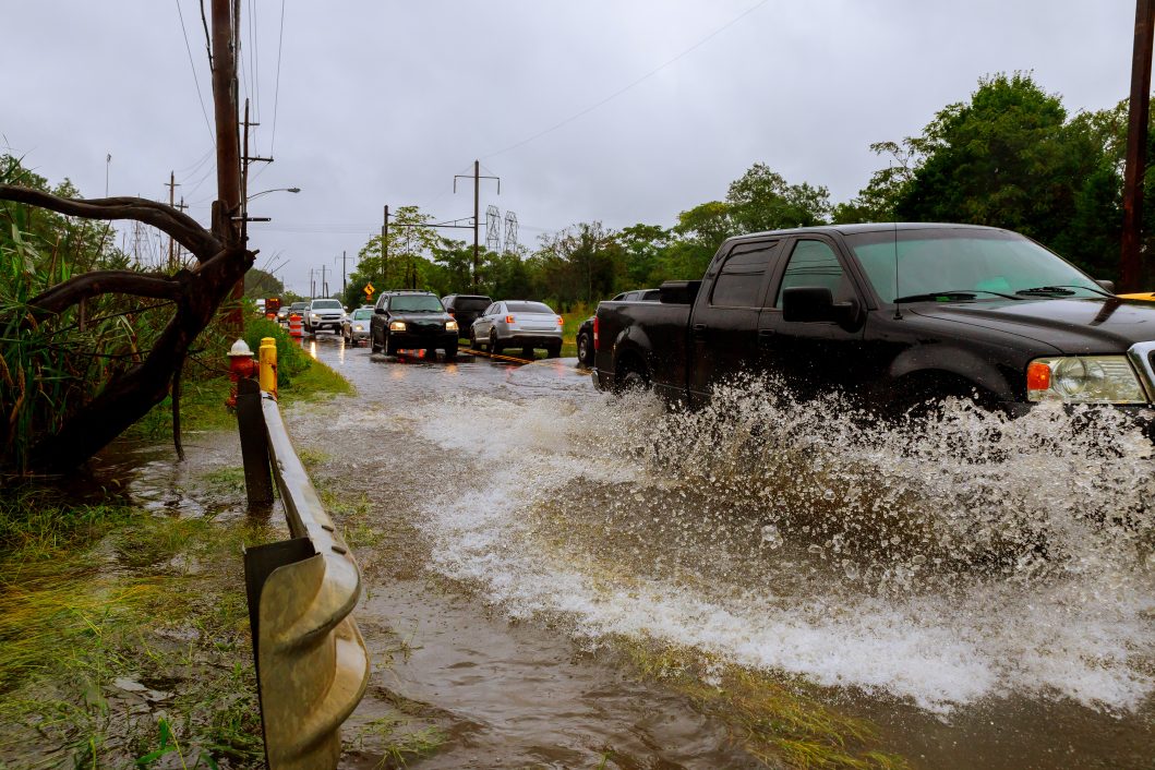 Black pickup truck driving through flooded road, water splashing, alongside trees and power lines.