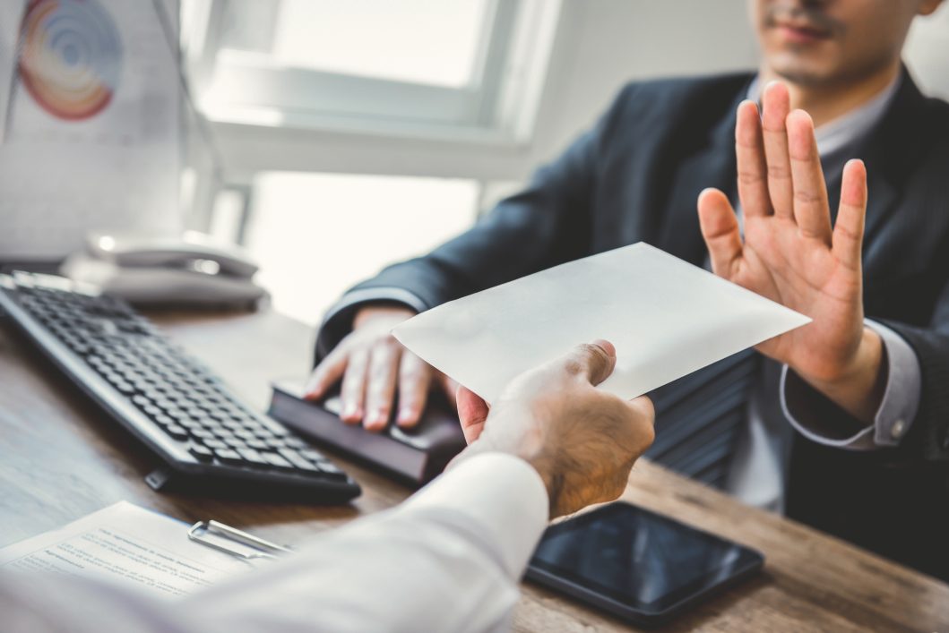 Person in suit refusing envelope, gesturing no with hand, sitting at office desk.