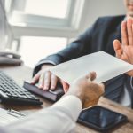 Person in suit refusing envelope, gesturing no with hand, sitting at office desk.