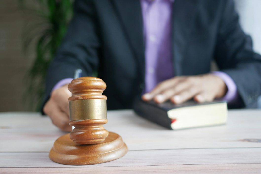 Wooden gavel on desk, person wearing suit resting hand on book in background.