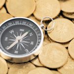 Compass resting on pile of gold coins, displaying cardinal directions on black and white dial