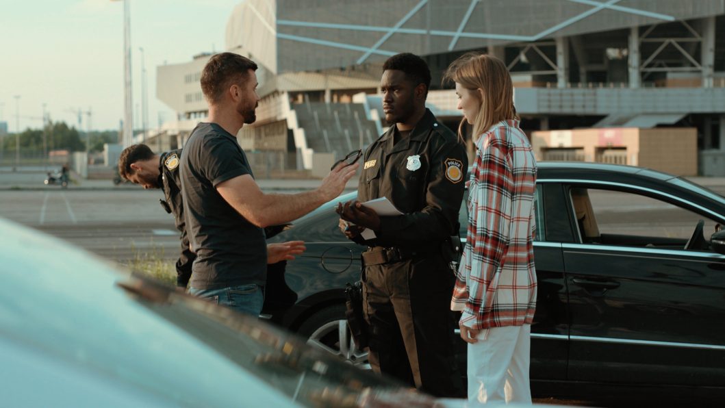 Man talking to police officers near black car outdoors in an open parking area.