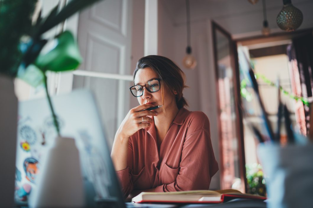 Person in glasses deep in thought, sitting at desk with laptop and open notebook.