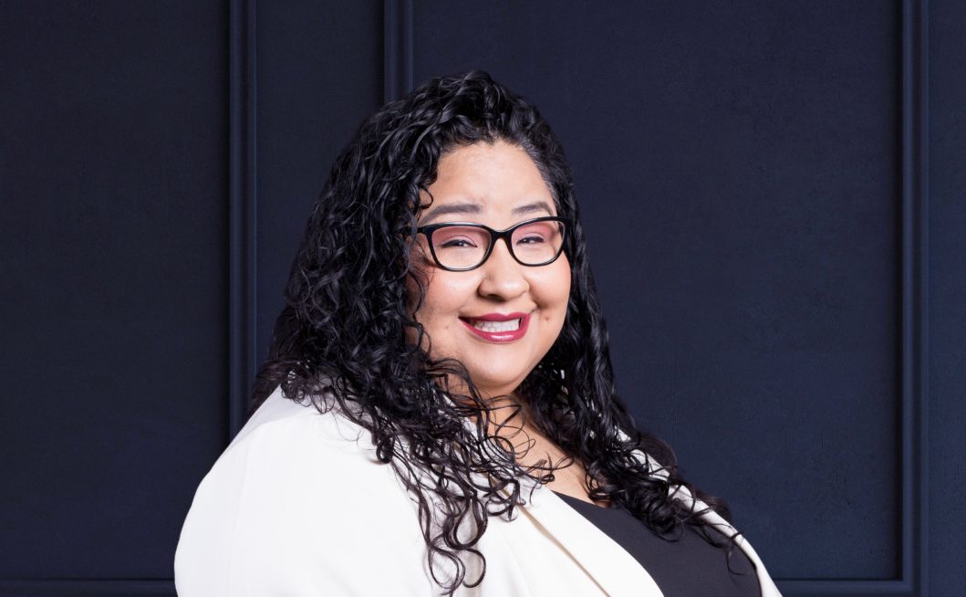 Smiling woman with curly hair and glasses, wearing white jacket against dark background.