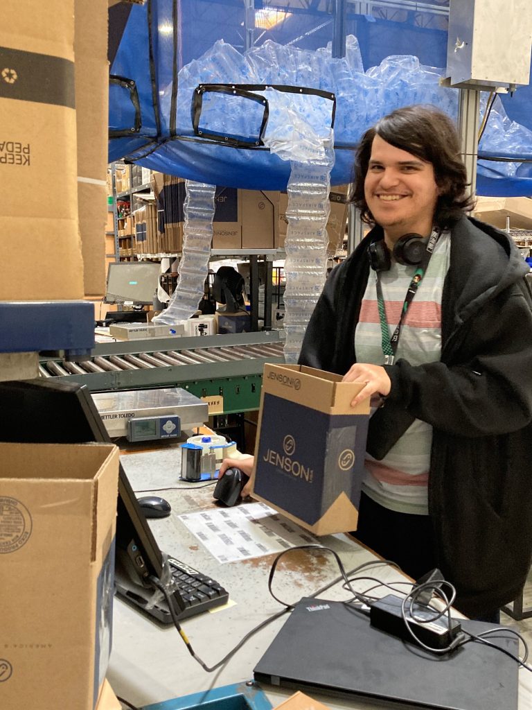 A smiling person standing at a desk with headphones around their neck assemble a package in a warehouse