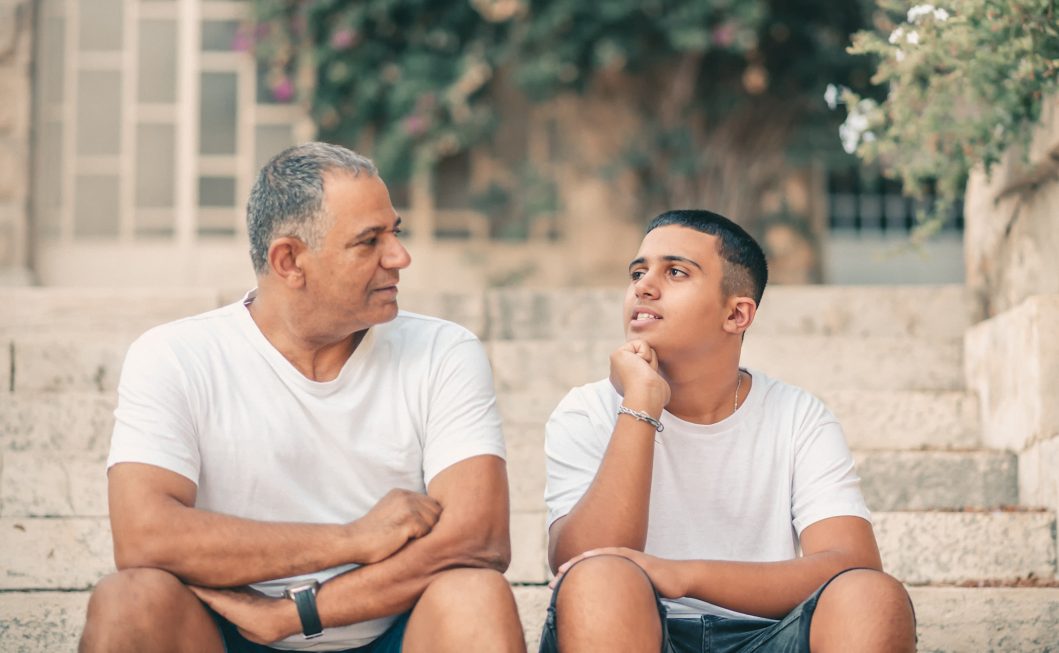 Two people sitting on outdoor stone steps, both wearing white shirts, looking at each other.