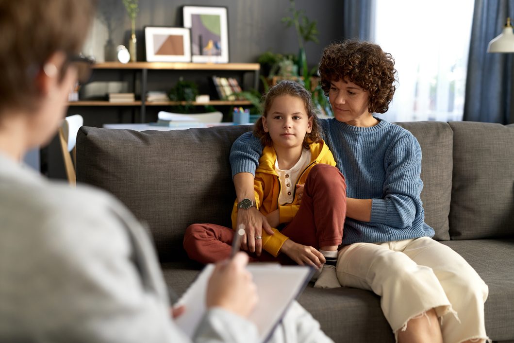 Therapist taking notes while mother and daughter sit on couch, both looking attentive and thoughtful.