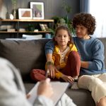 Therapist taking notes while mother and daughter sit on couch, both looking attentive and thoughtful.