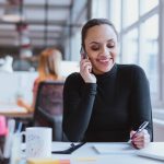 Person smiling while talking on phone, holding pen, seated at desk with office supplies.
