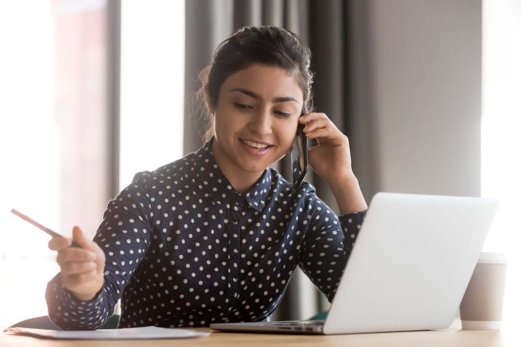 Woman in polka dot blouse talking on phone, holding pen, working on laptop at desk.