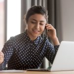 Woman in polka dot blouse talking on phone, holding pen, working on laptop at desk.