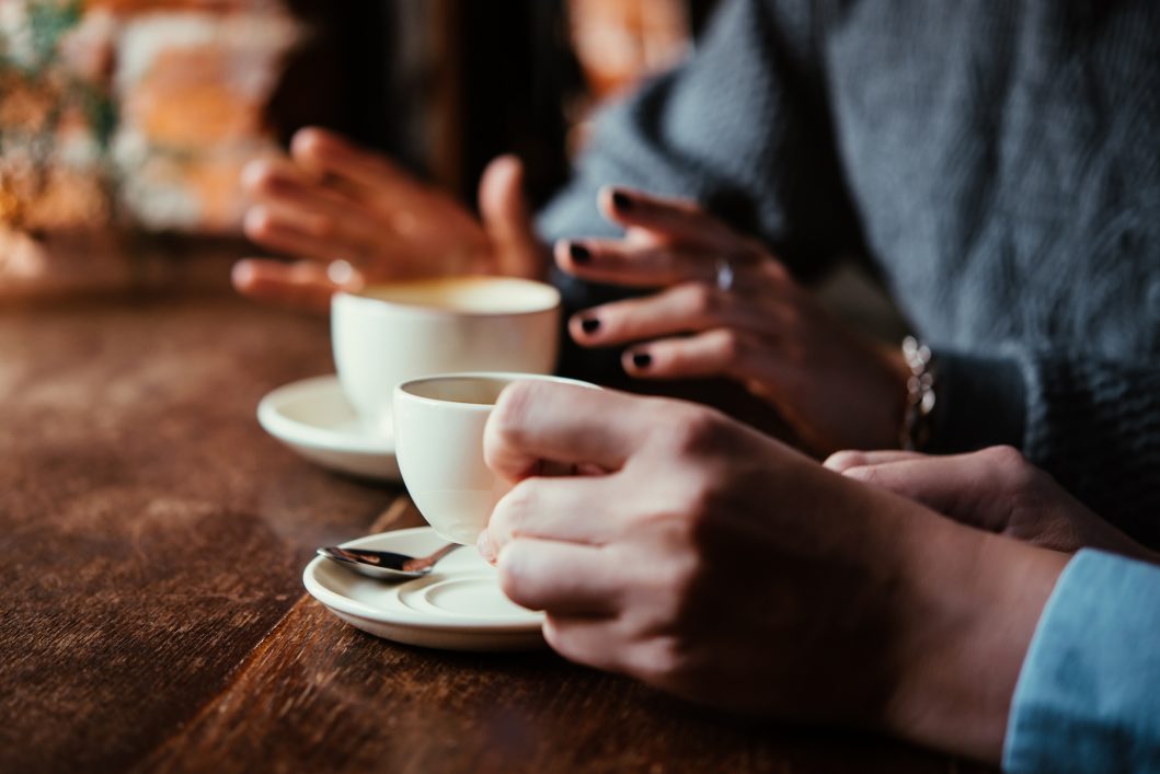 Hands holding cups of coffee on wooden table, engaged in conversation.