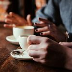 Hands holding cups of coffee on wooden table, engaged in conversation.