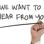 Hand holding pen writing "We Want to Hear From You" on white background.