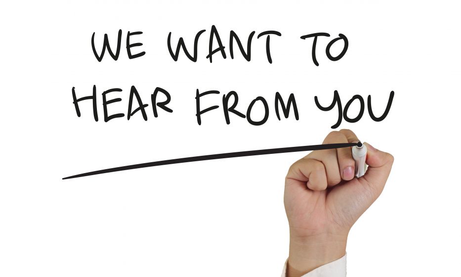 Hand holding pen writing "We Want to Hear From You" on white background.