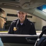Police officer smiling at driver through open car window, building in background.