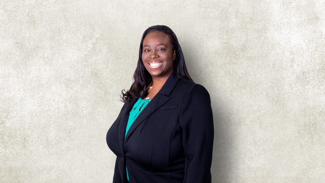 Smiling woman in black blazer and green top against textured gray background.