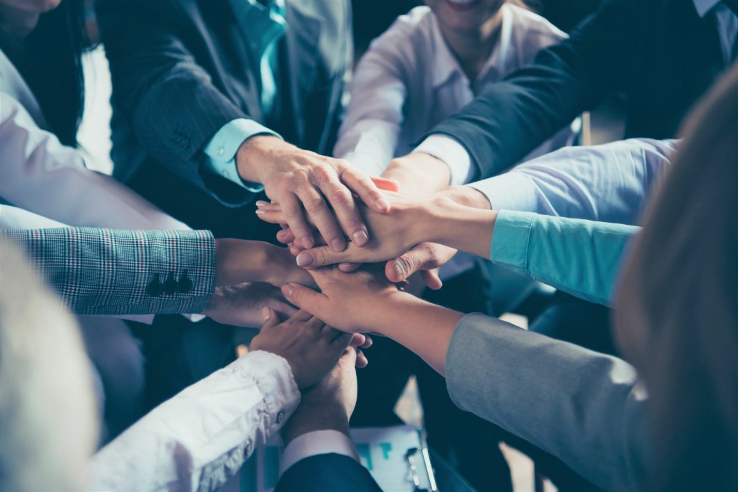 Group of people stacking hands in center, showing teamwork and collaboration, wearing business attire.