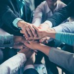 Group of people stacking hands in center, showing teamwork and collaboration, wearing business attire.