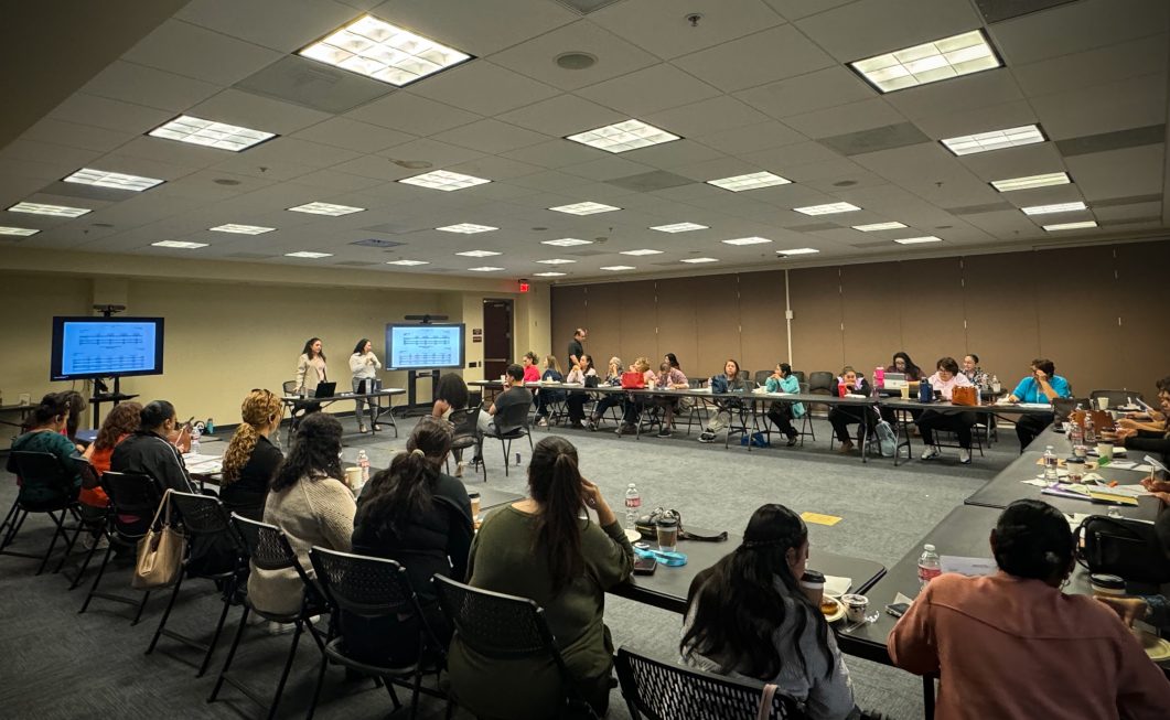 People sitting around large conference table, two women presenting, screens displaying information, bright room.