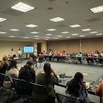 People sitting around large conference table, two women presenting, screens displaying information, bright room.