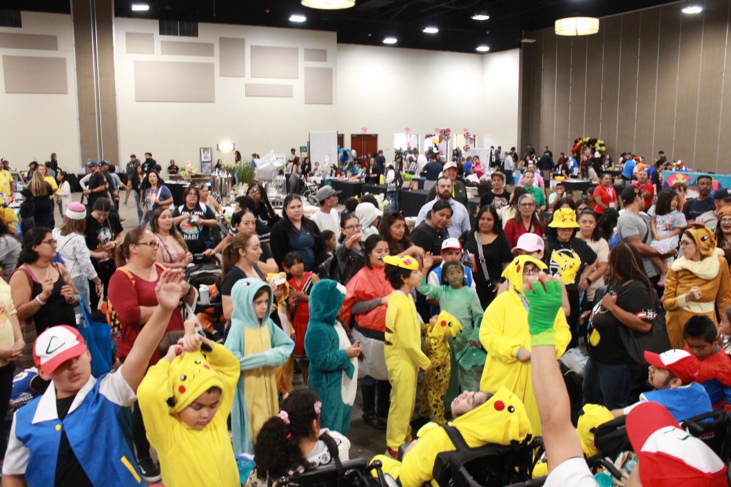 Crowded indoor event with children and adults wearing colorful Pokémon costumes, many Pikachu outfits visible.