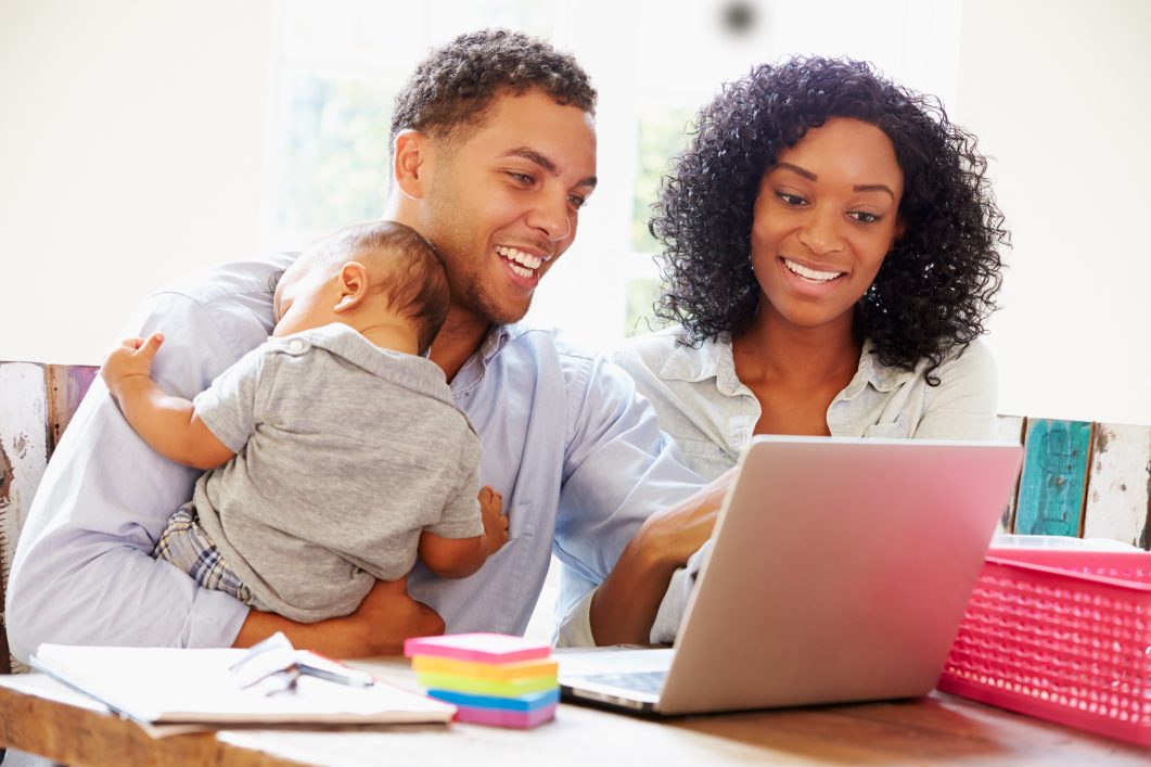Father holding baby and mother smiling at laptop, sitting at table with colorful papers.