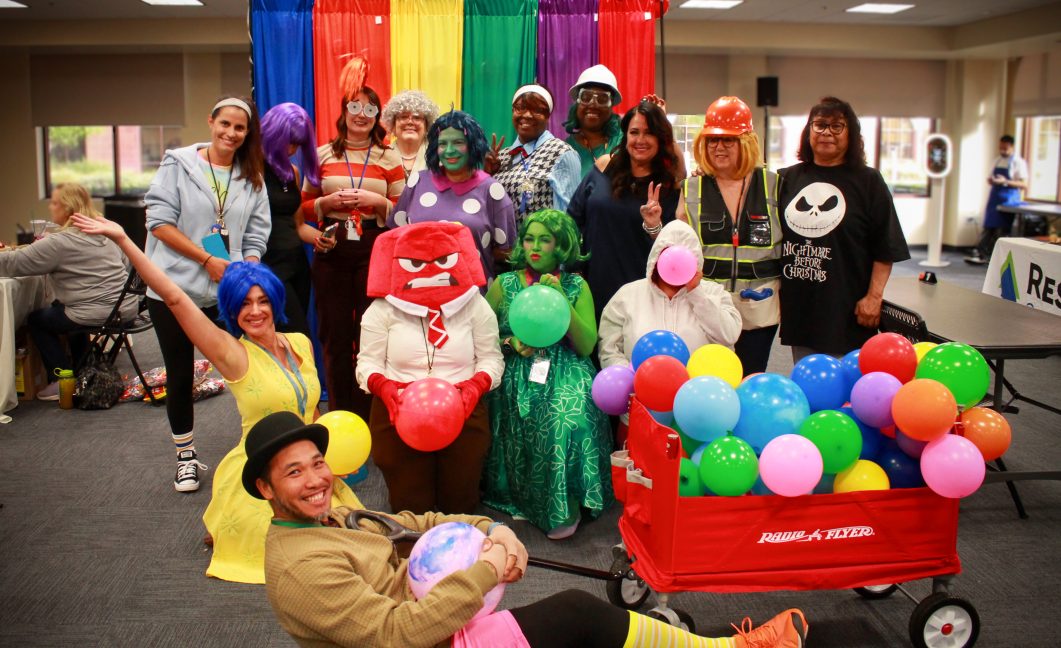 Group of people in colorful costumes and wigs with balloons, posing indoors, red wagon in front.