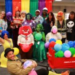 Group of people in colorful costumes and wigs with balloons, posing indoors, red wagon in front.