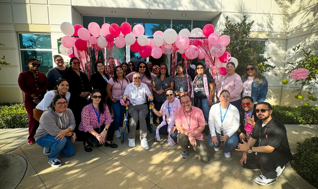 Group of people wearing pink gathered outside, surrounded by pink and white balloons, smiling together.