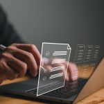 Hands holding stylus touching virtual checklist on open laptop with digital documents.